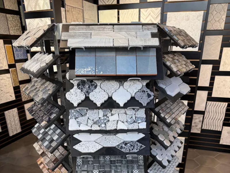Showroom display of stone and tile samples: a central stacked rack with patterned mosaic pieces, hexagonal and irregular shapes, and surrounding shelves filled with square and rectangular tile swatches.